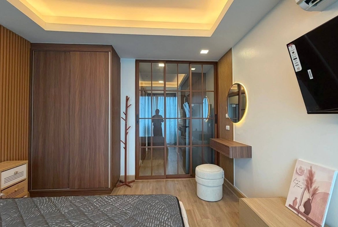 Hotel-style bedroom with wooden wardrobe, glass sliding door, round backlit mirror, and wall-mounted TV to the right; bed in foreground.