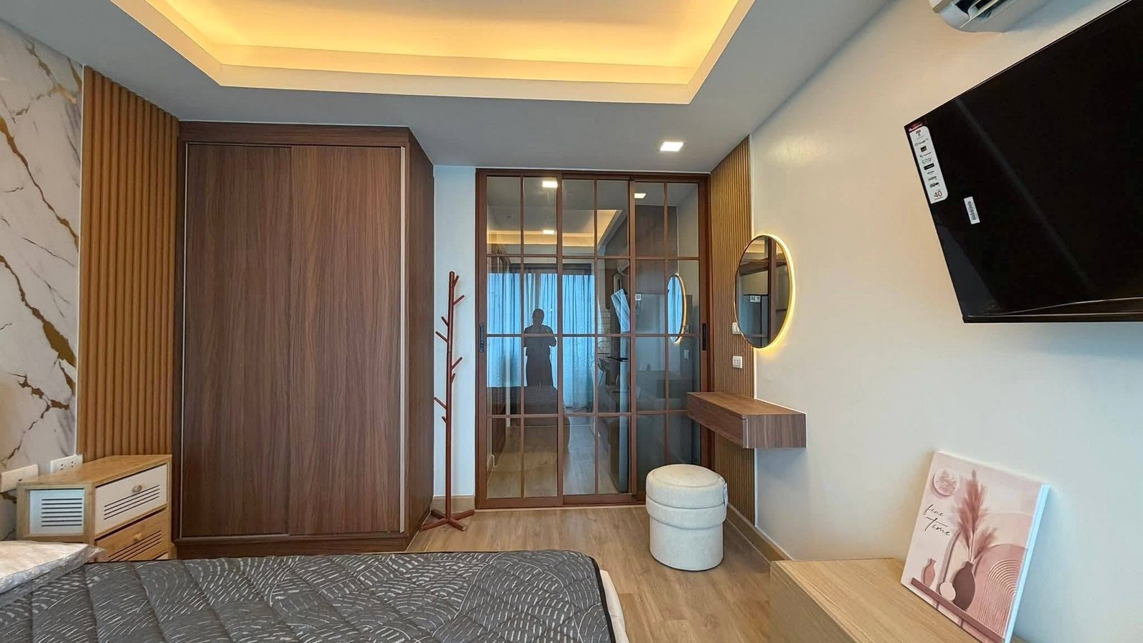 LBR2_Pattaya_Sale_Dlc613_6 Hotel-style bedroom with wooden wardrobe, glass sliding door, round backlit mirror, and wall-mounted TV to the right; bed in foreground.
