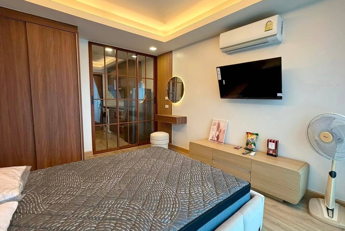 Bedroom with a large wooden wardrobe, glass sliding partition, and a bed with a gray patterned comforter in the foreground.