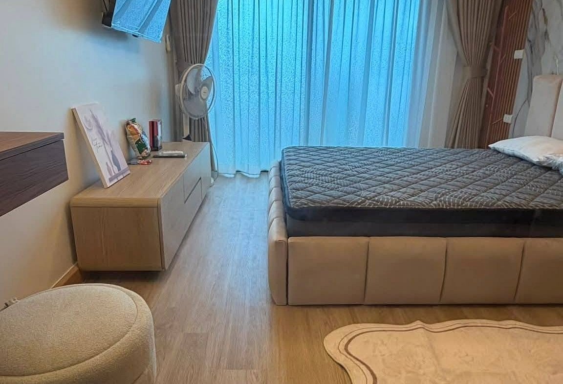 Hotel bedroom with a low platform bed, gray quilt, wall-mounted TV, and large window with sheer curtains.