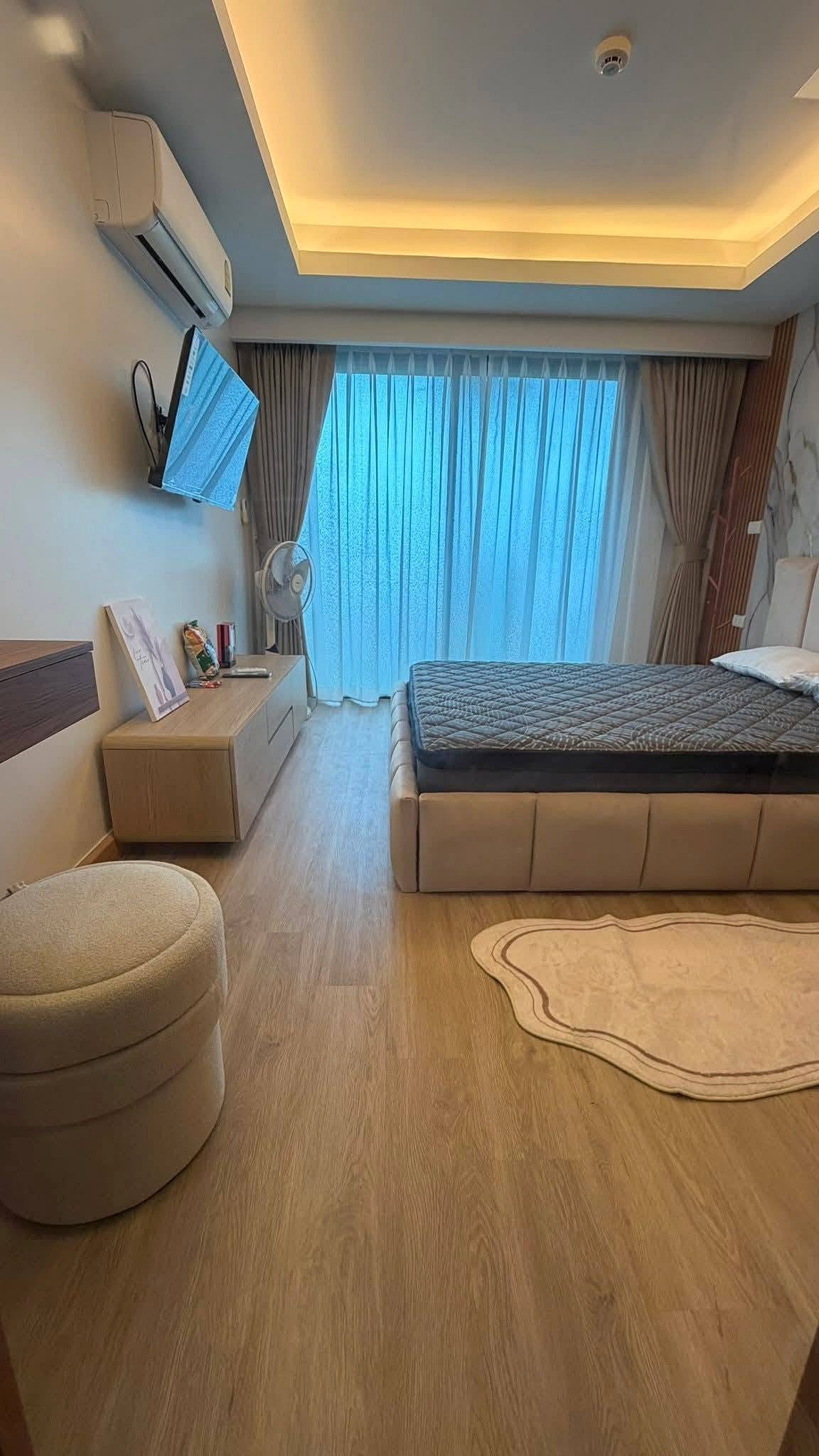 LBR2_Pattaya_Sale_Dlc613_9 Hotel bedroom with a low platform bed, gray quilt, wall-mounted TV, and large window with sheer curtains.