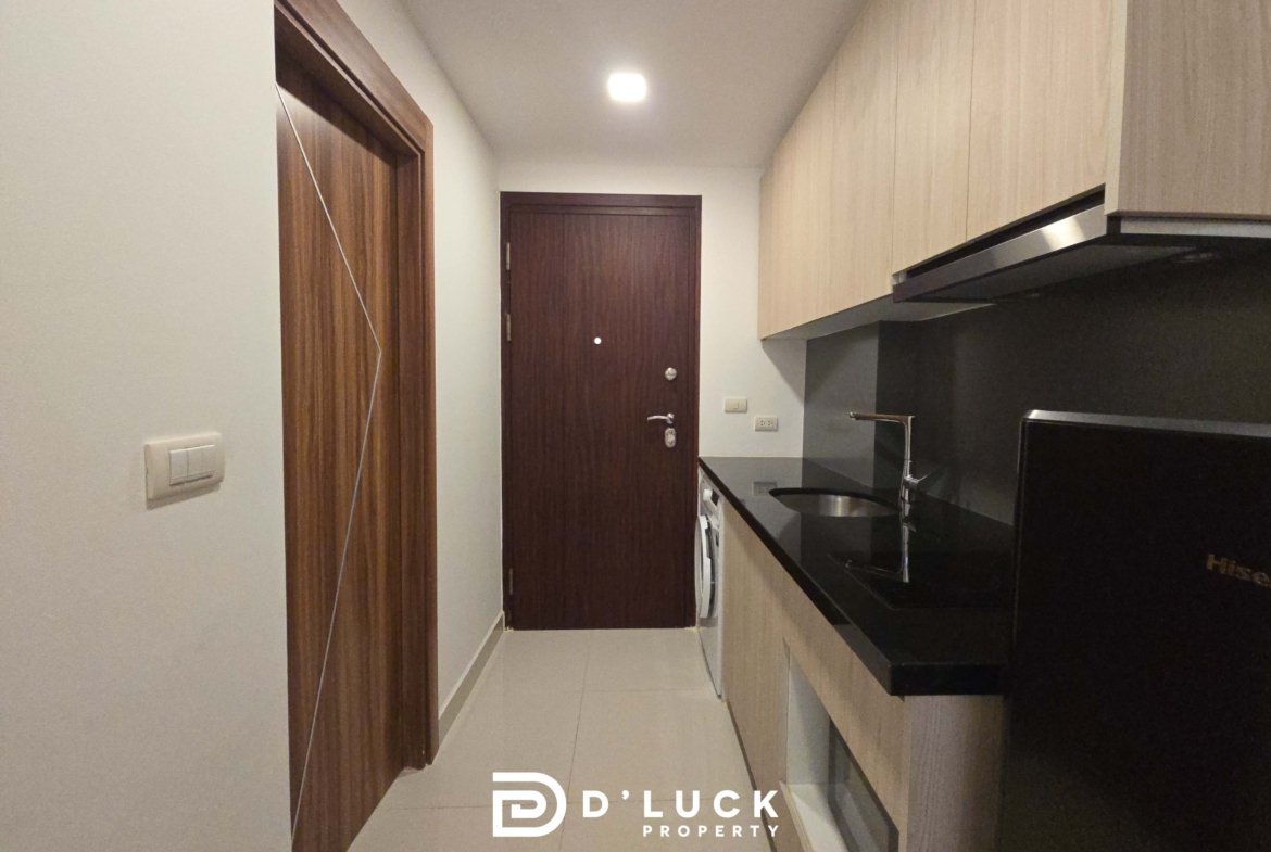 Narrow kitchen hallway with light wood cabinets on the right, a dark countertop, and a dark wooden door at the end.