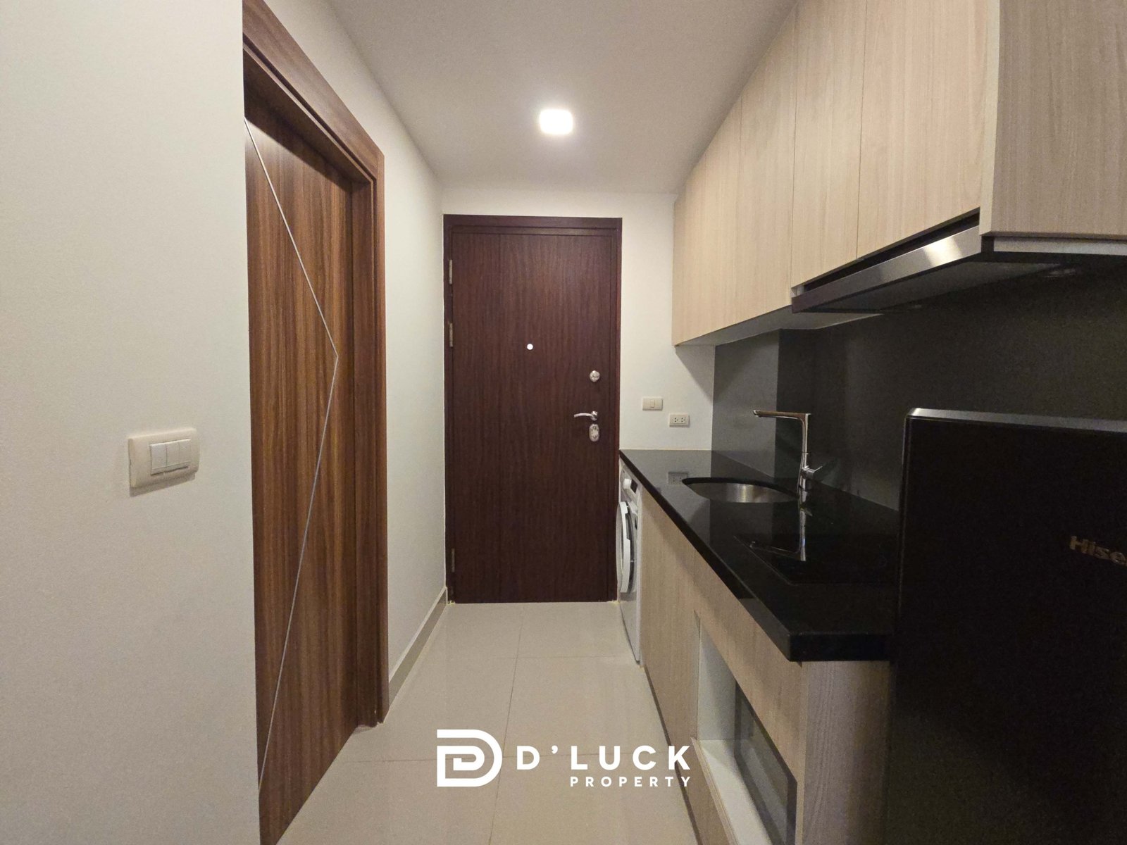 Narrow kitchen hallway with light wood cabinets on the right, a dark countertop, and a dark wooden door at the end.