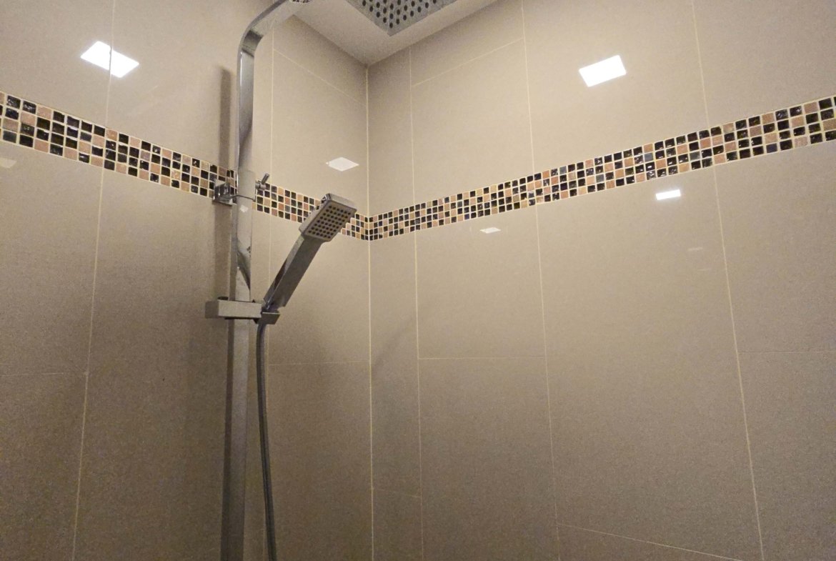 Modern shower with a square ceiling rain showerhead and a handheld wand in a beige tiled enclosure with a mosaic wall border (D’LUCK Property watermark).