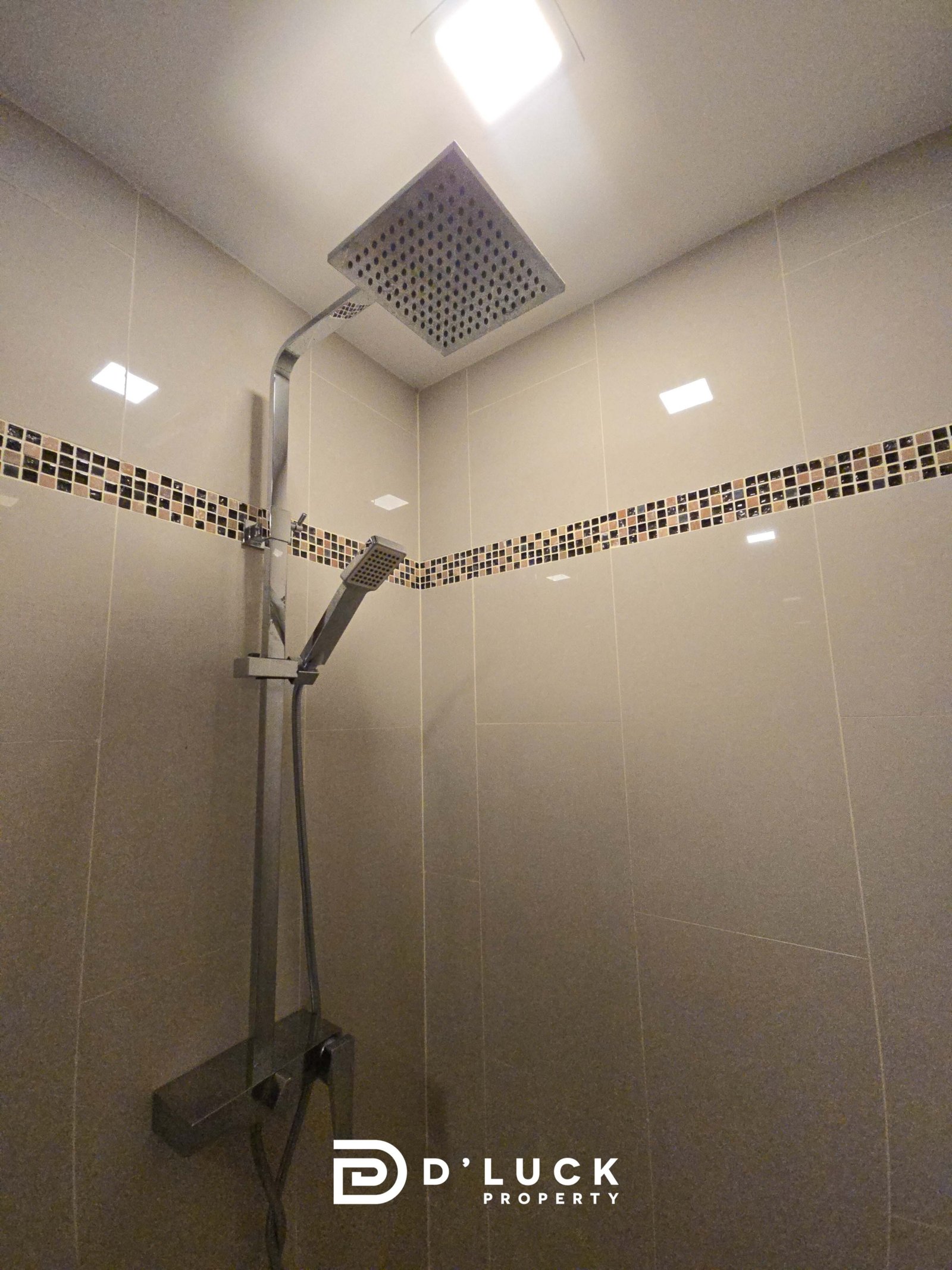 Modern shower with a square ceiling rain showerhead and a handheld wand in a beige tiled enclosure with a mosaic wall border (D’LUCK Property watermark).