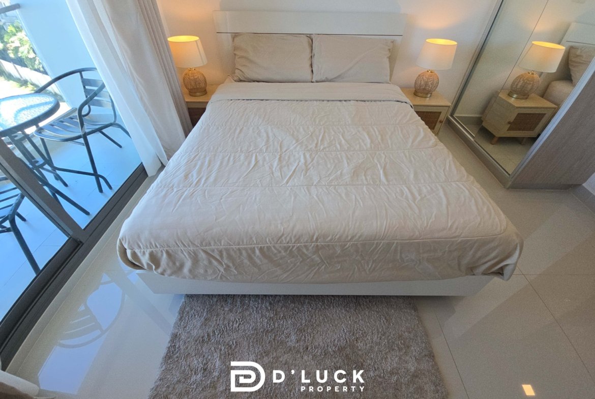 Bright, minimalist hotel bedroom with a white bed, two beige pillows, and wicker-lamp nightstands; glass balcony door on the left.