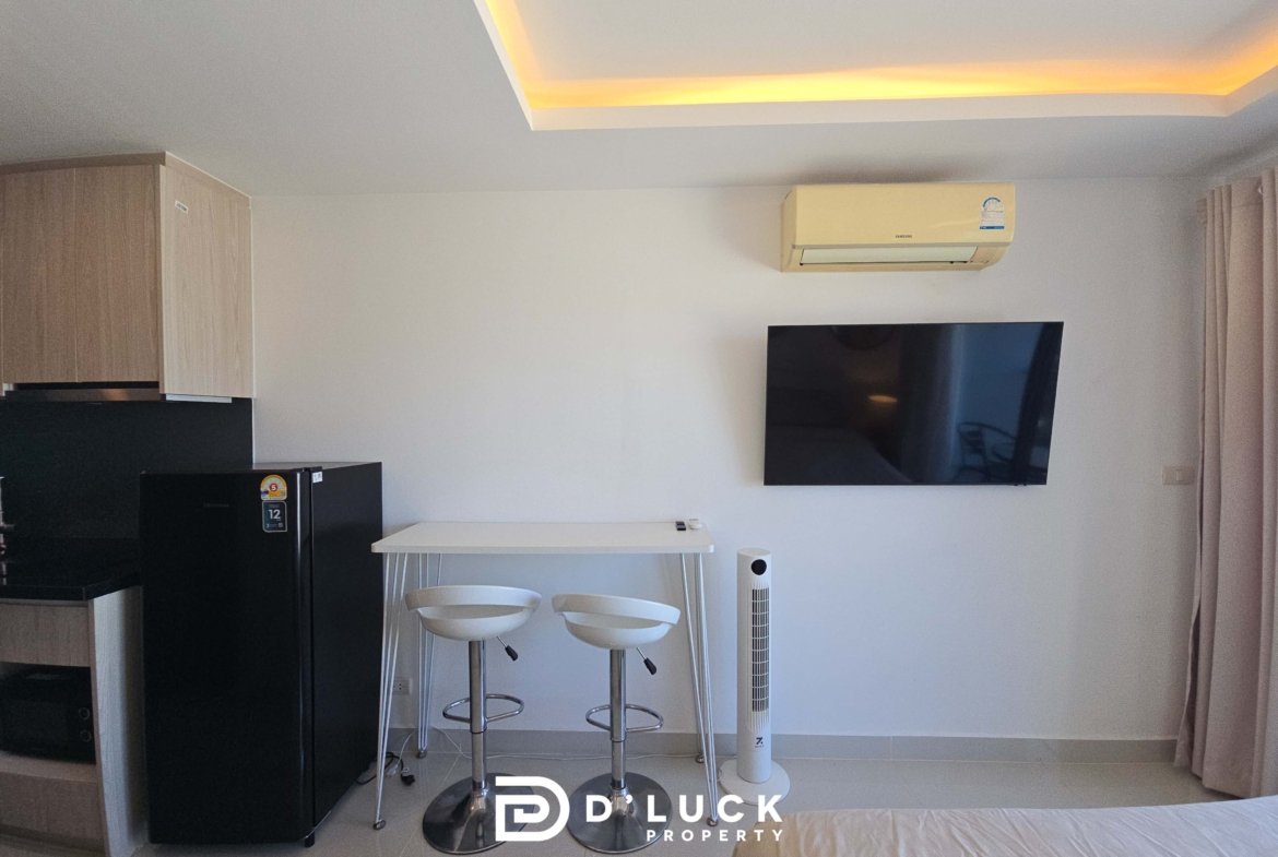 Modern studio living area with a wall-mounted TV, beige air conditioner, black kitchen fridge, and a white table with two tall white stools near light curtains.