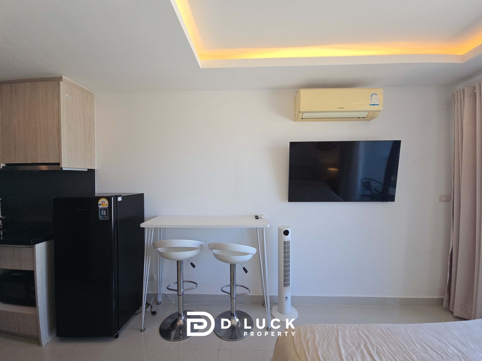 Modern studio living area with a wall-mounted TV, beige air conditioner, black kitchen fridge, and a white table with two tall white stools near light curtains.