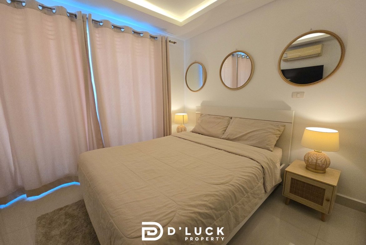 Bedroom with two beige beds, matching nightstands, wicker lamps, and beige curtains under soft ambient lighting. Three round mirrors above the headboard wall.