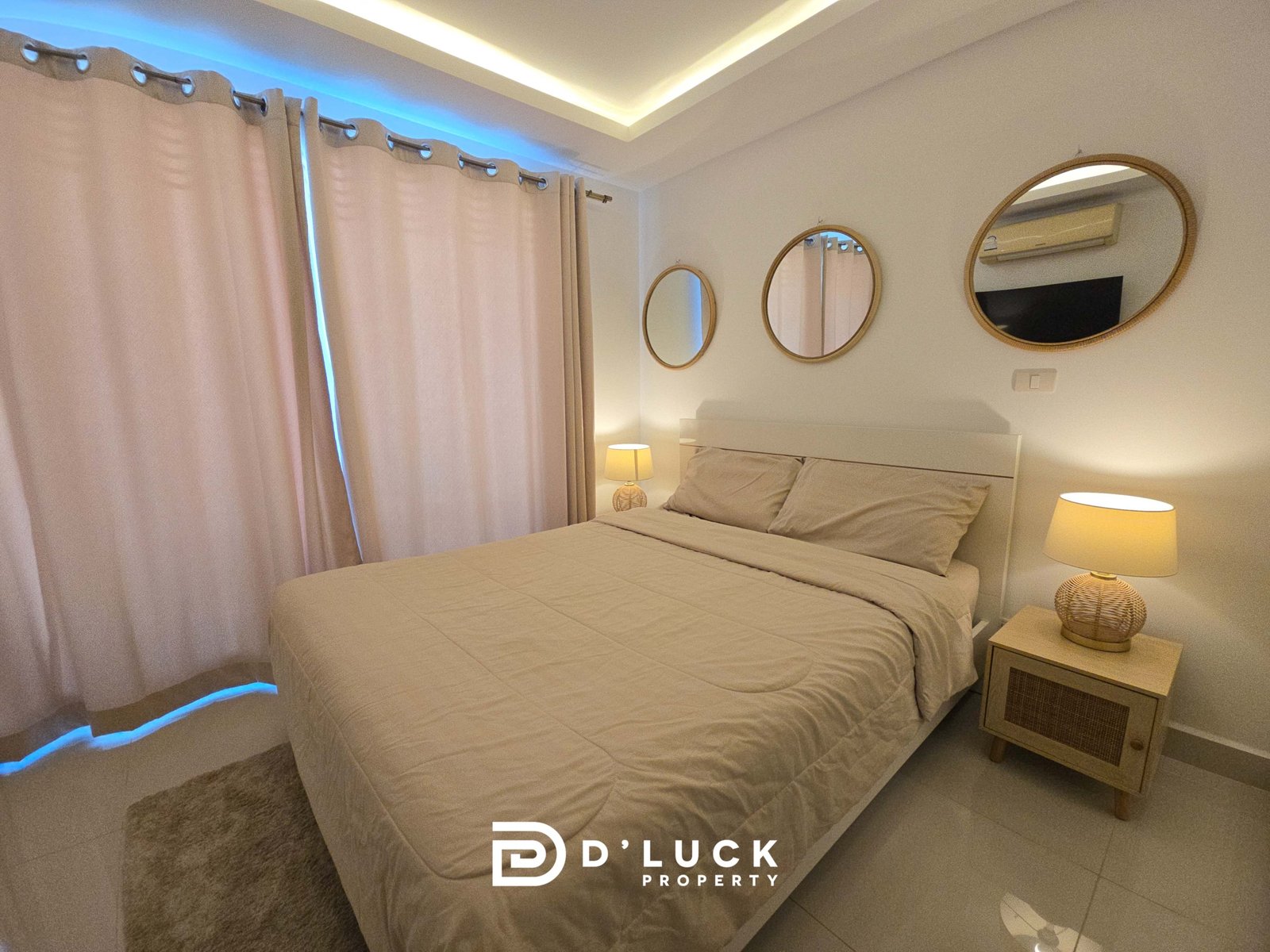 Bedroom with two beige beds, matching nightstands, wicker lamps, and beige curtains under soft ambient lighting. Three round mirrors above the headboard wall.