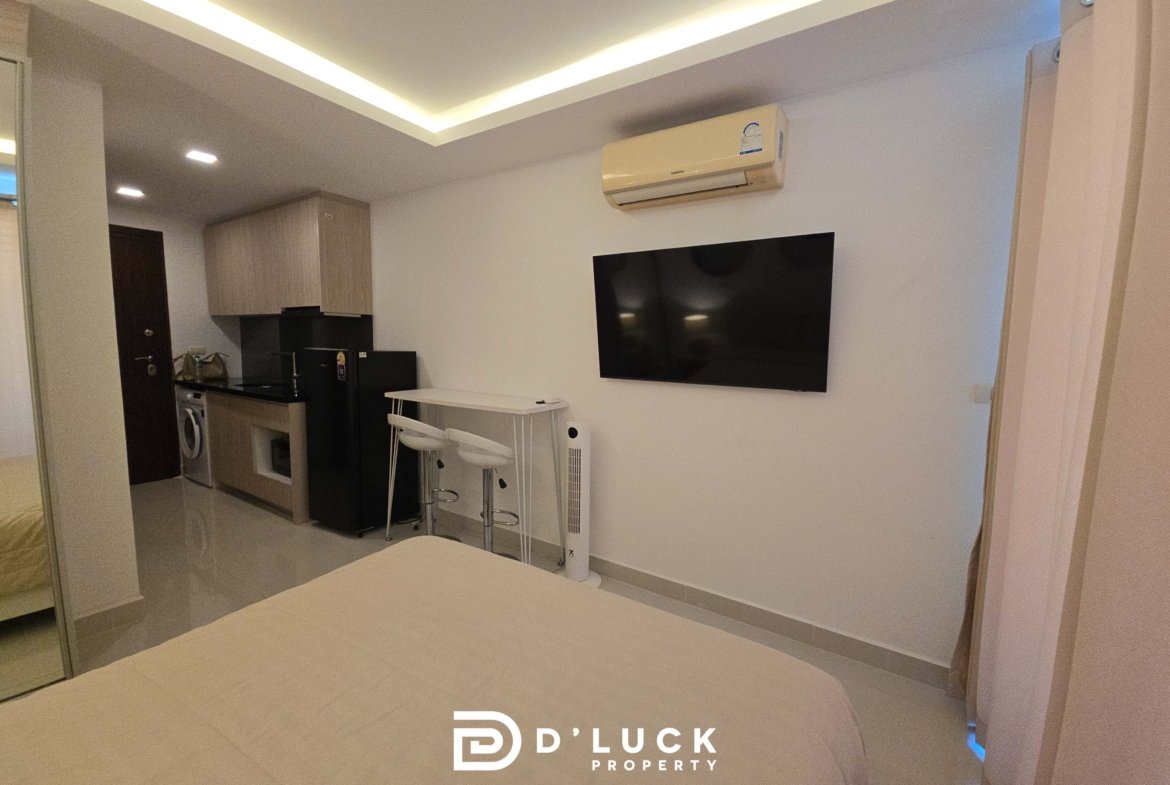 Modern living area with wall-mounted TV, ceiling LED coves, and a compact kitchen featuring light wood cabinets and a black countertop.