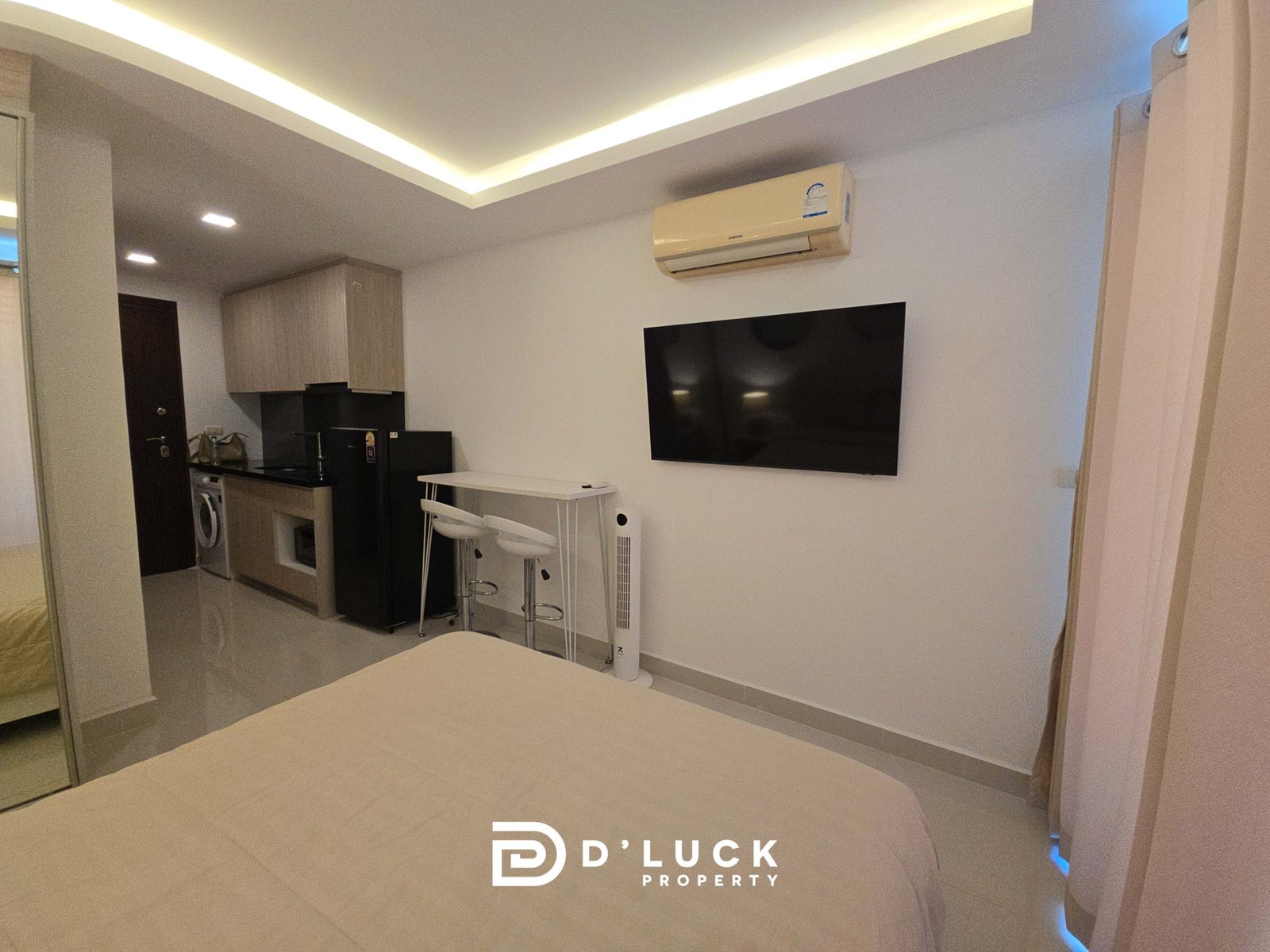 Modern living area with wall-mounted TV, ceiling LED coves, and a compact kitchen featuring light wood cabinets and a black countertop.