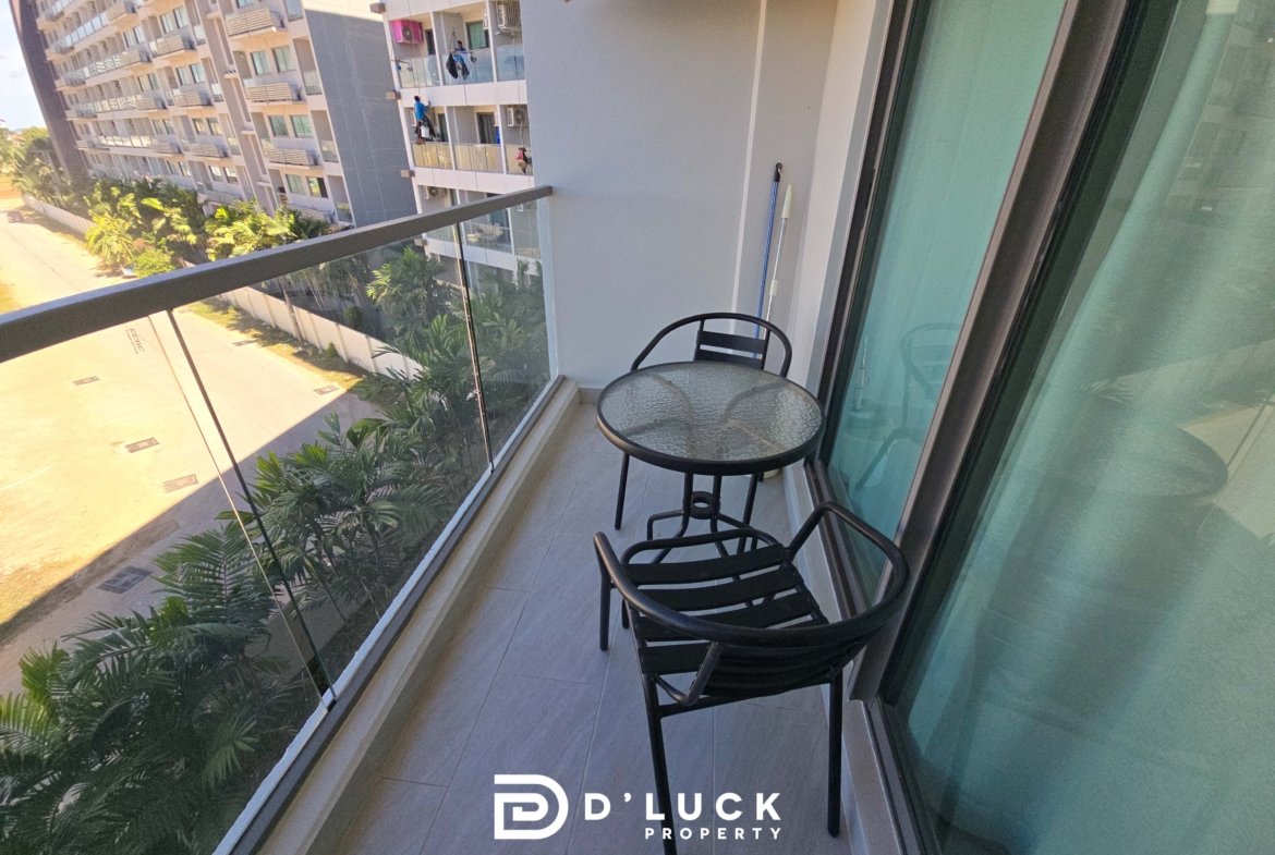 Balcony with glass railing, two black metal chairs and a glass table, overlooking modern apartment buildings and palm trees; D’LUCK Property logo.