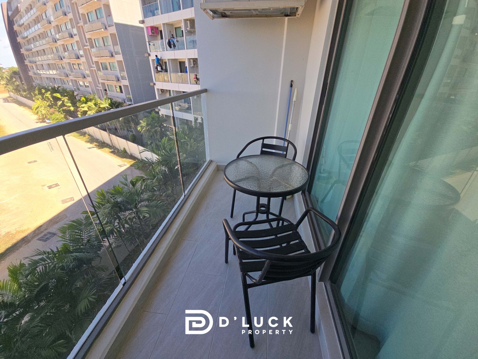 Balcony with glass railing, two black metal chairs and a glass table, overlooking modern apartment buildings and palm trees; D’LUCK Property logo.