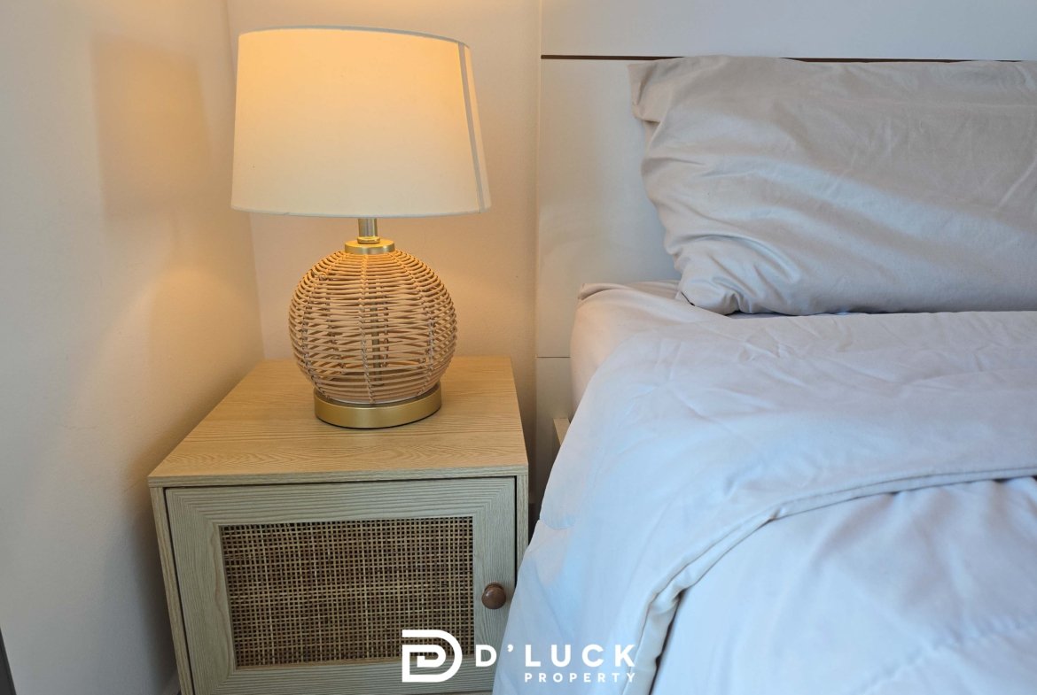 Bedside table with a round wicker lamp beside a neatly made bed in a bright bedroom (watermark reads Duck Property)./n