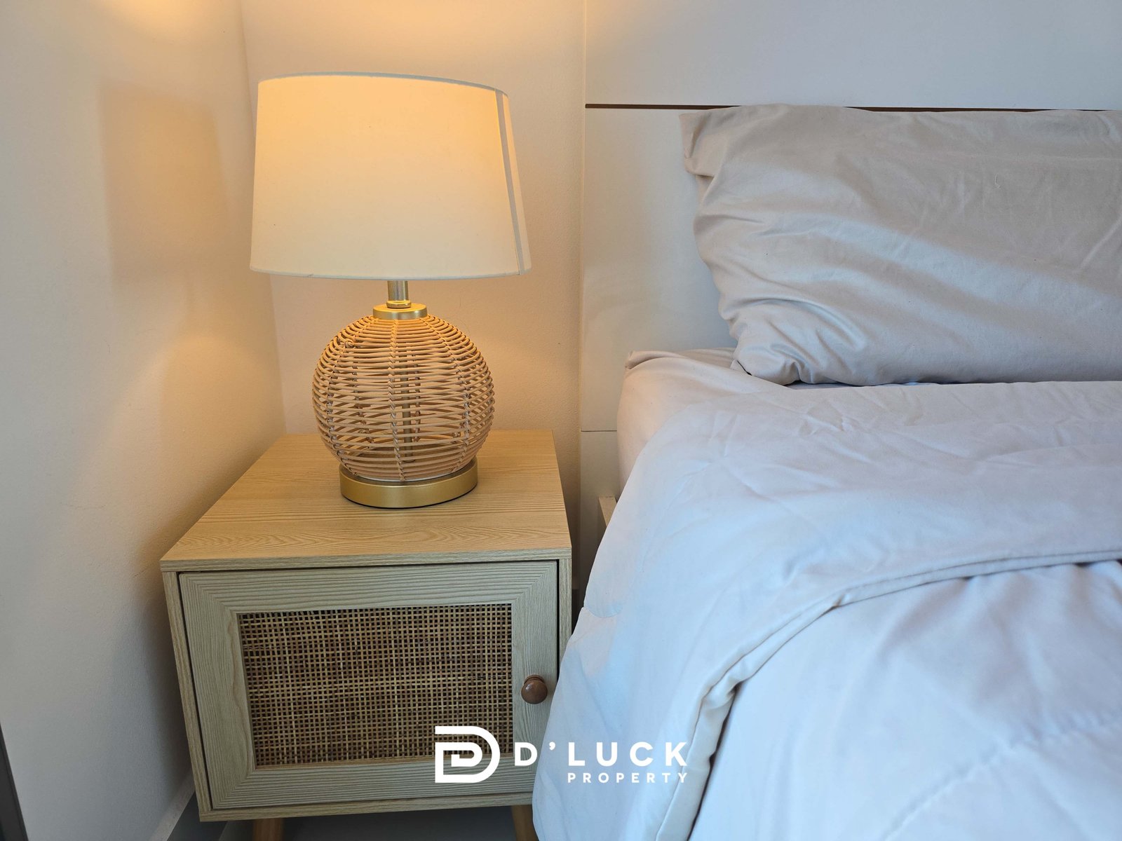 Bedside table with a round wicker lamp beside a neatly made bed in a bright bedroom (watermark reads Duck Property)./n