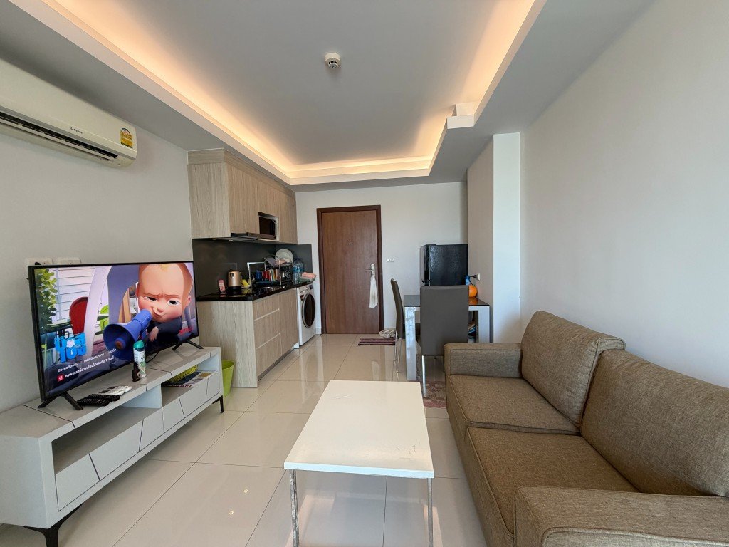 Open-plan living area with a beige sofa facing a TV, white coffee table, and a compact kitchen; an air conditioner is on the wall above the TV.