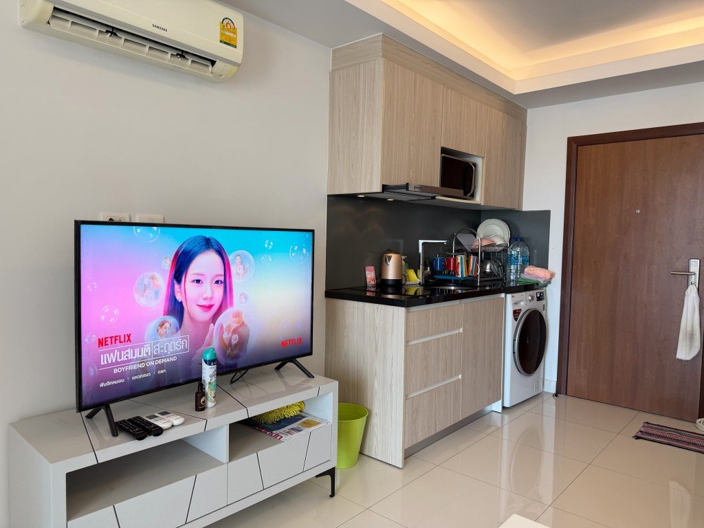 Compact living area with a TV on a white stand beside a small kitchenette and doorway to the right, plus an air conditioner above.