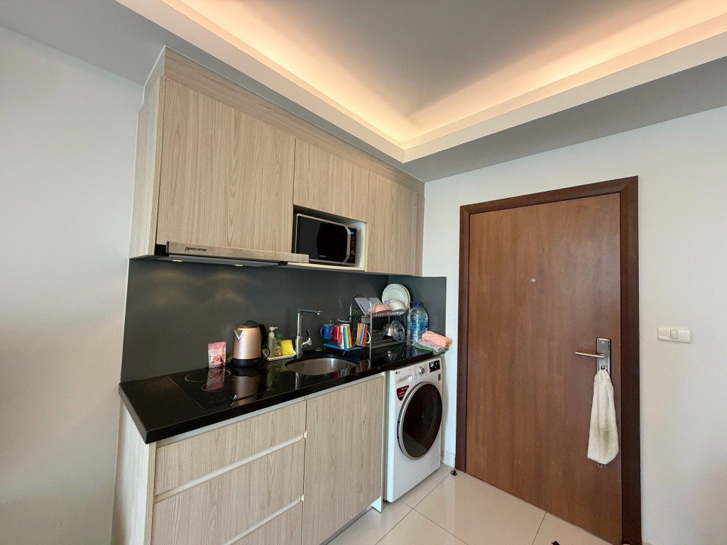 Compact kitchen with light wood cabinets, black countertop, microwave, kettle, and a front-loading washing machine beside a wooden door.