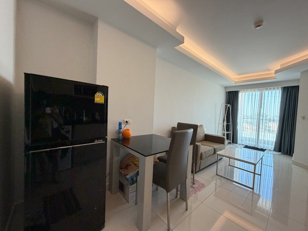 Open-plan living room with a black refrigerator, glass dining table, gray chairs, beige sofa, and a balcony with sheer curtains.