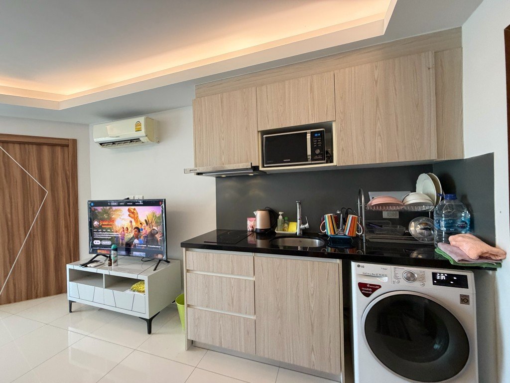 Open-plan kitchen and living area with light wood cabinets, black countertop, sink, drying rack, washing machine, and a TV on a stand beyond.