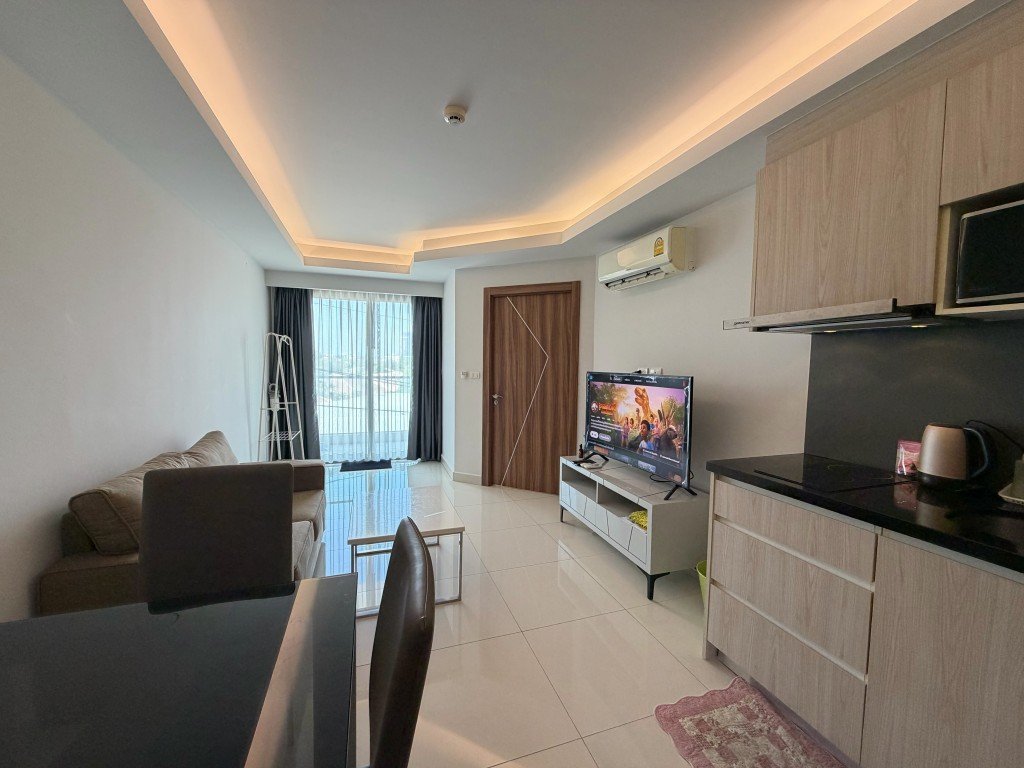 Open-concept living room with a beige sofa facing a TV on a white media console and a balcony door with sheer curtains in the background.