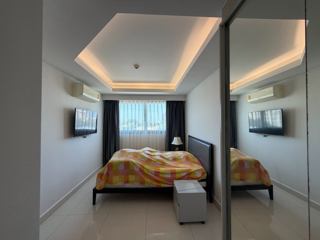 Bright modern bedroom with a double bed, gray headboard, and a colorful checkered duvet, plus a wall-mounted TV and ceiling-cove lighting.