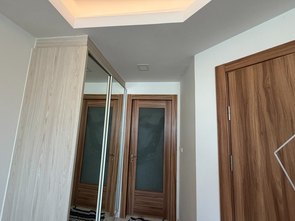 Modern hallway with light wood doors and a tall mirrored wardrobe on the left, reflecting a wooden door with frosted glass panels on the opposite wall.