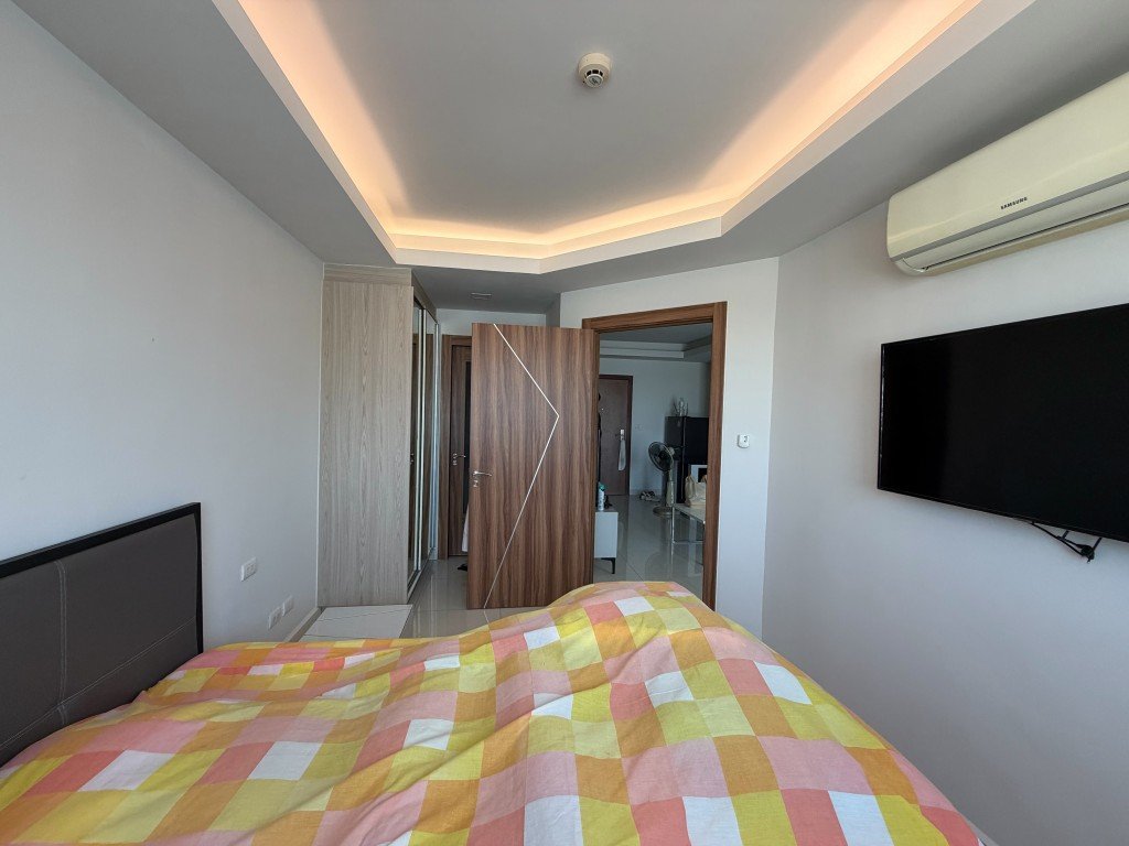 Bedroom with a bed covered by a yellow–pink checkered quilt, a wall-mounted TV, and ceiling with warm recessed lighting; open door to a hallway.
