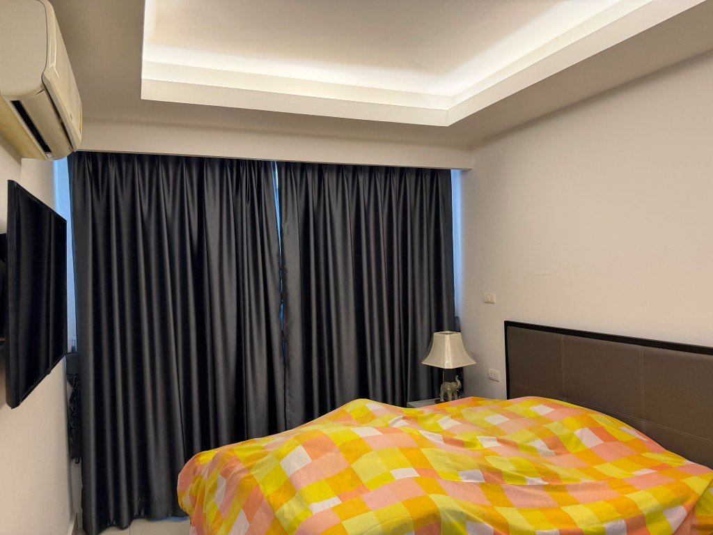 Bedroom with a colorful yellow-pink plaid duvet, dark curtains, wall-mounted TV, air conditioner, and bedside lamp on a nightstand.