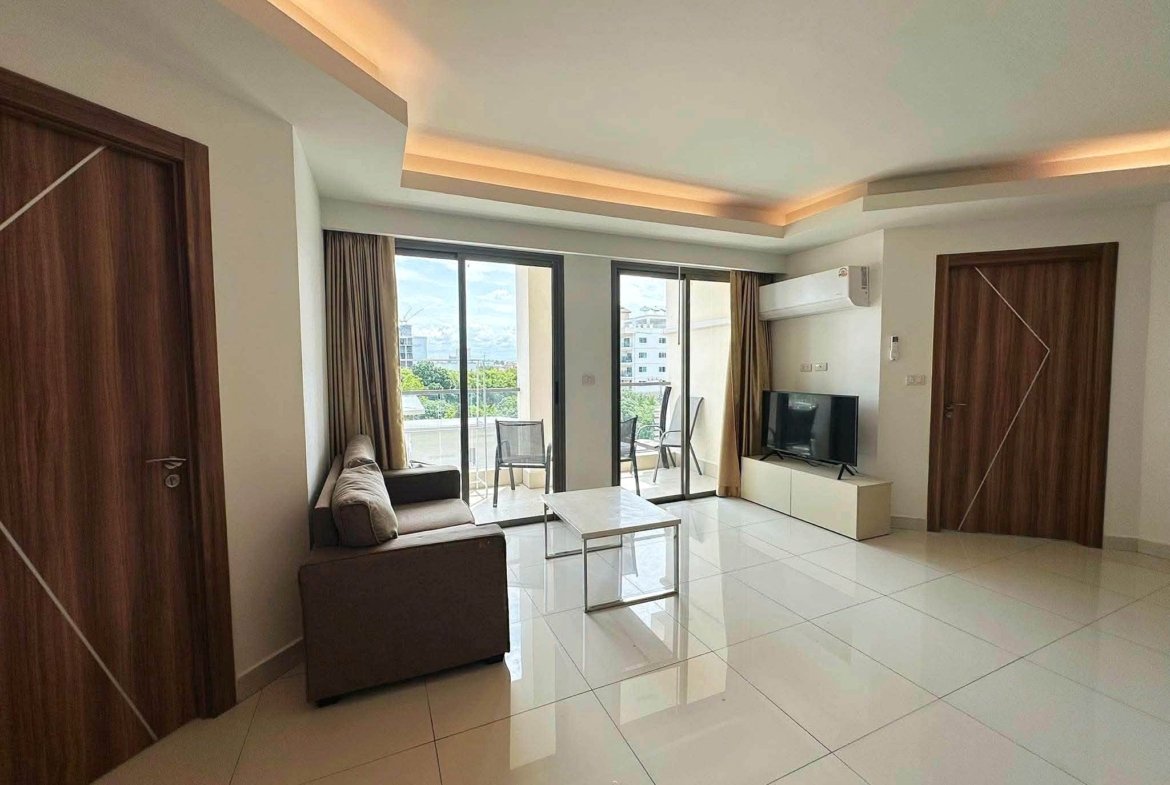 Living room with white glossy tile floor, brown wooden doors, and a sofa facing a TV unit; sliding glass doors open to a balcony with a city/view outside.
