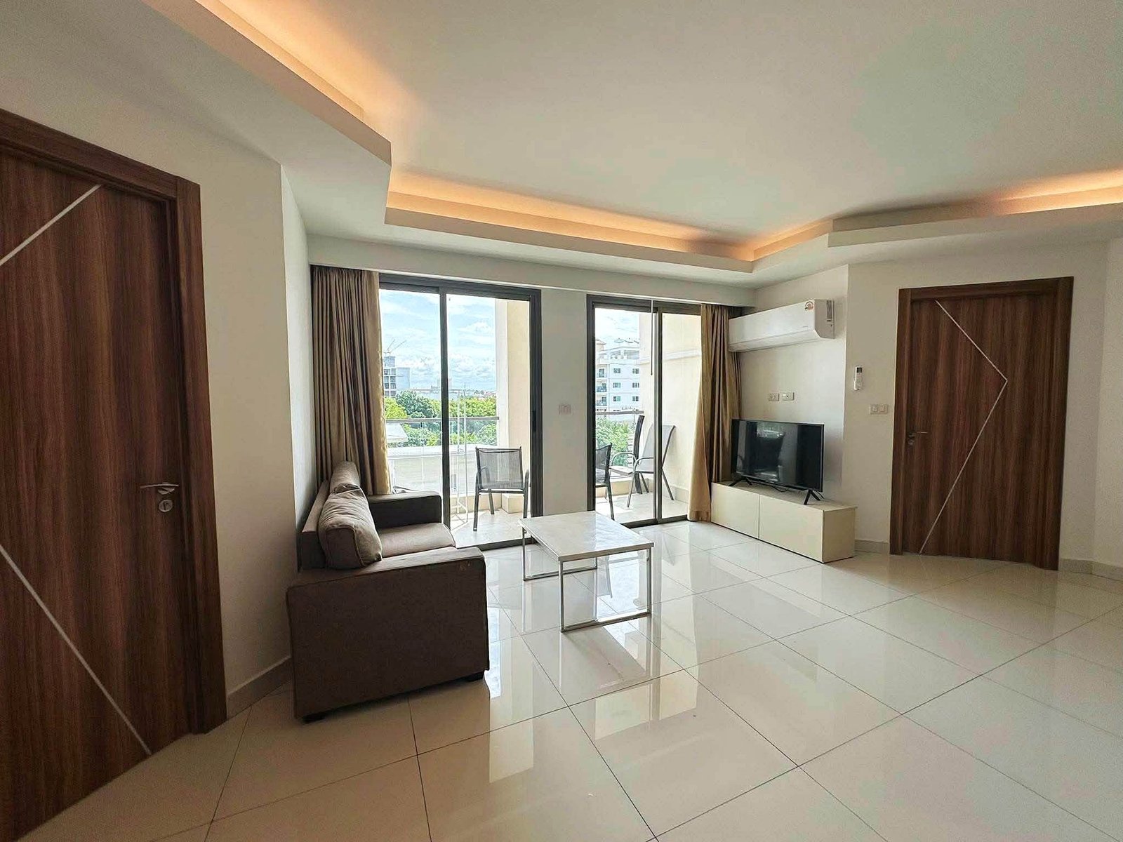 Laguna_2_Pattaya_Sale_Dlc636 Living room with white glossy tile floor, brown wooden doors, and a sofa facing a TV unit; sliding glass doors open to a balcony with a city/view outside.