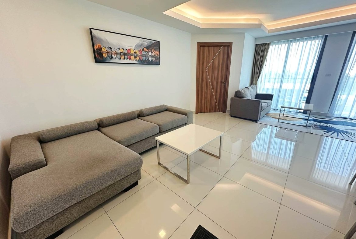 Modern living room with a gray sectional sofa, white coffee table, and a colorful village painting on the wall near a wooden door.