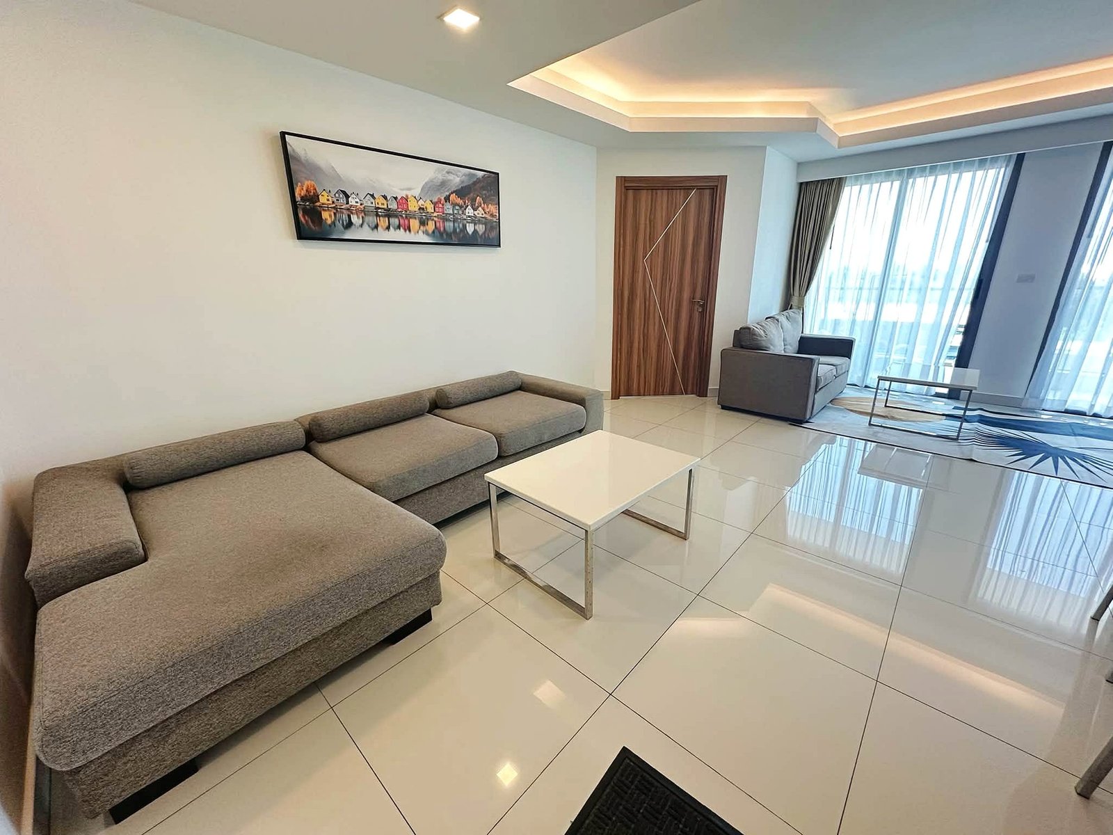 Laguna_2_Pattaya_Sale_Dlc636_2 Modern living room with a gray sectional sofa, white coffee table, and a colorful village painting on the wall near a wooden door.