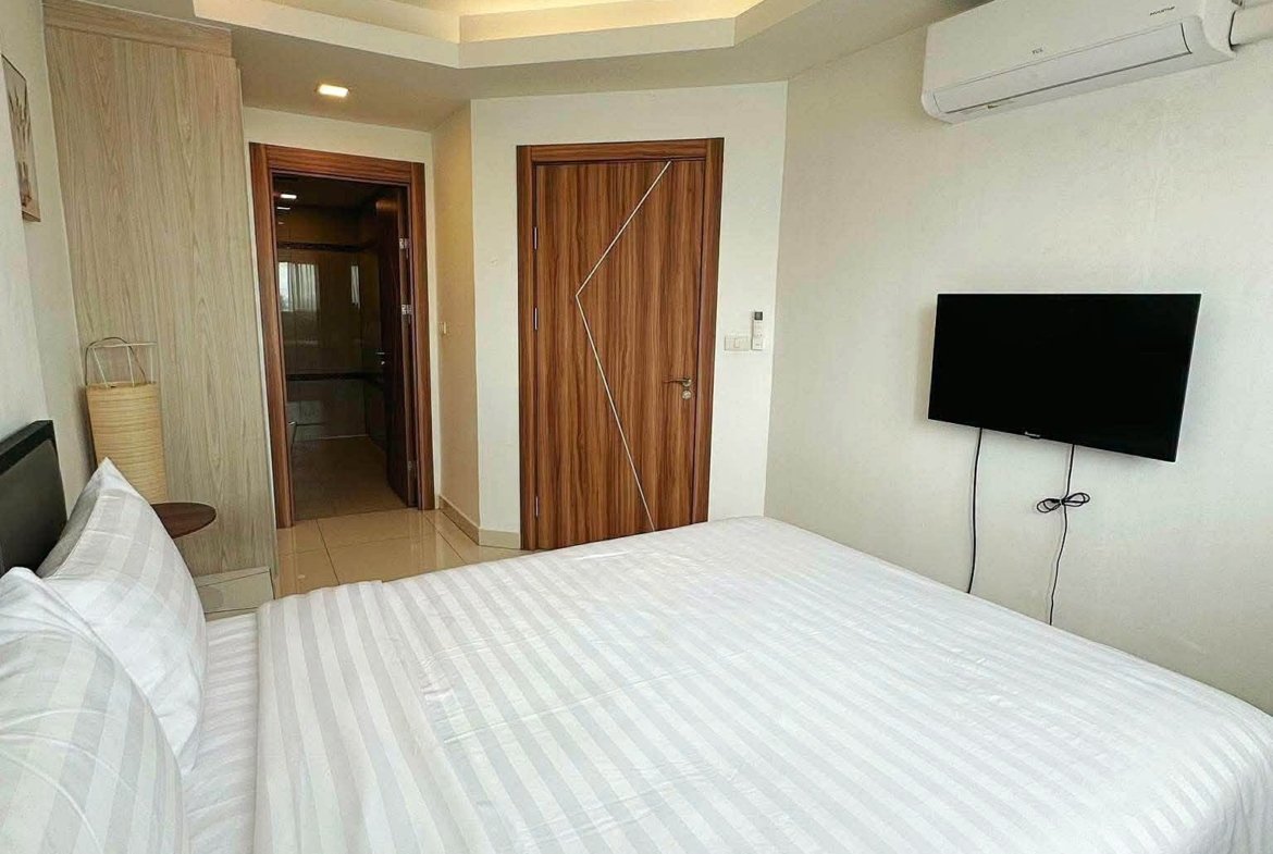 Hotel bedroom with white striped bedding, wall-mounted TV, and wooden doors under soft recessed ceiling lights.
