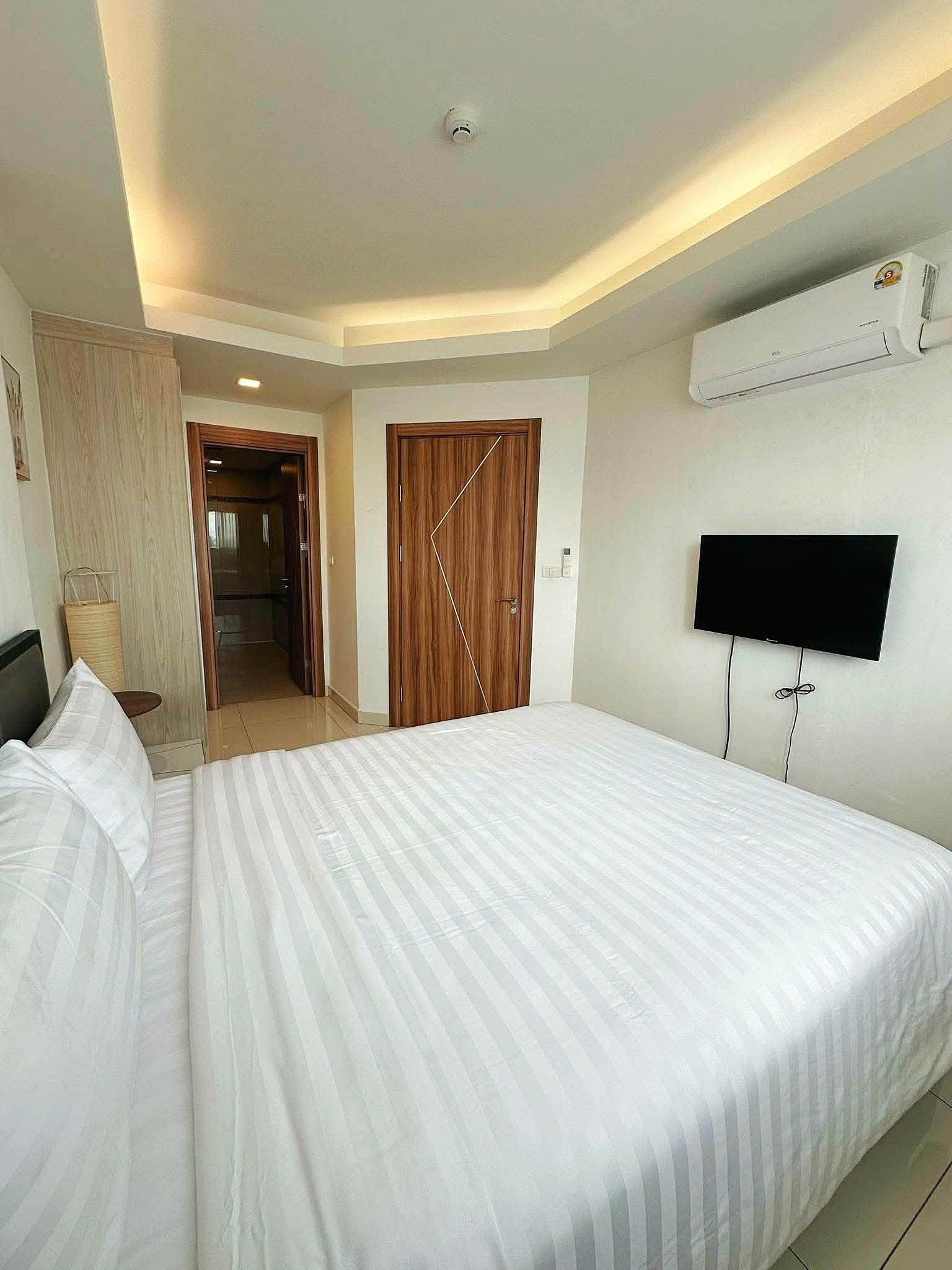 Laguna_2_Pattaya_Sale_Dlc636_3 Hotel bedroom with white striped bedding, wall-mounted TV, and wooden doors under soft recessed ceiling lights.