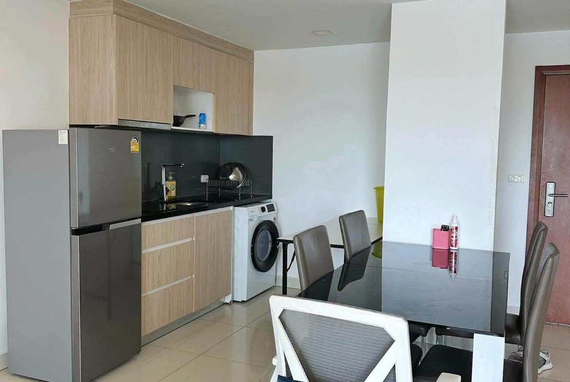 Modern kitchen and dining area with wooden cabinets, stainless steel fridge, washing machine, glass dining table, and gray chairs; white partition wall and tiled floor.
