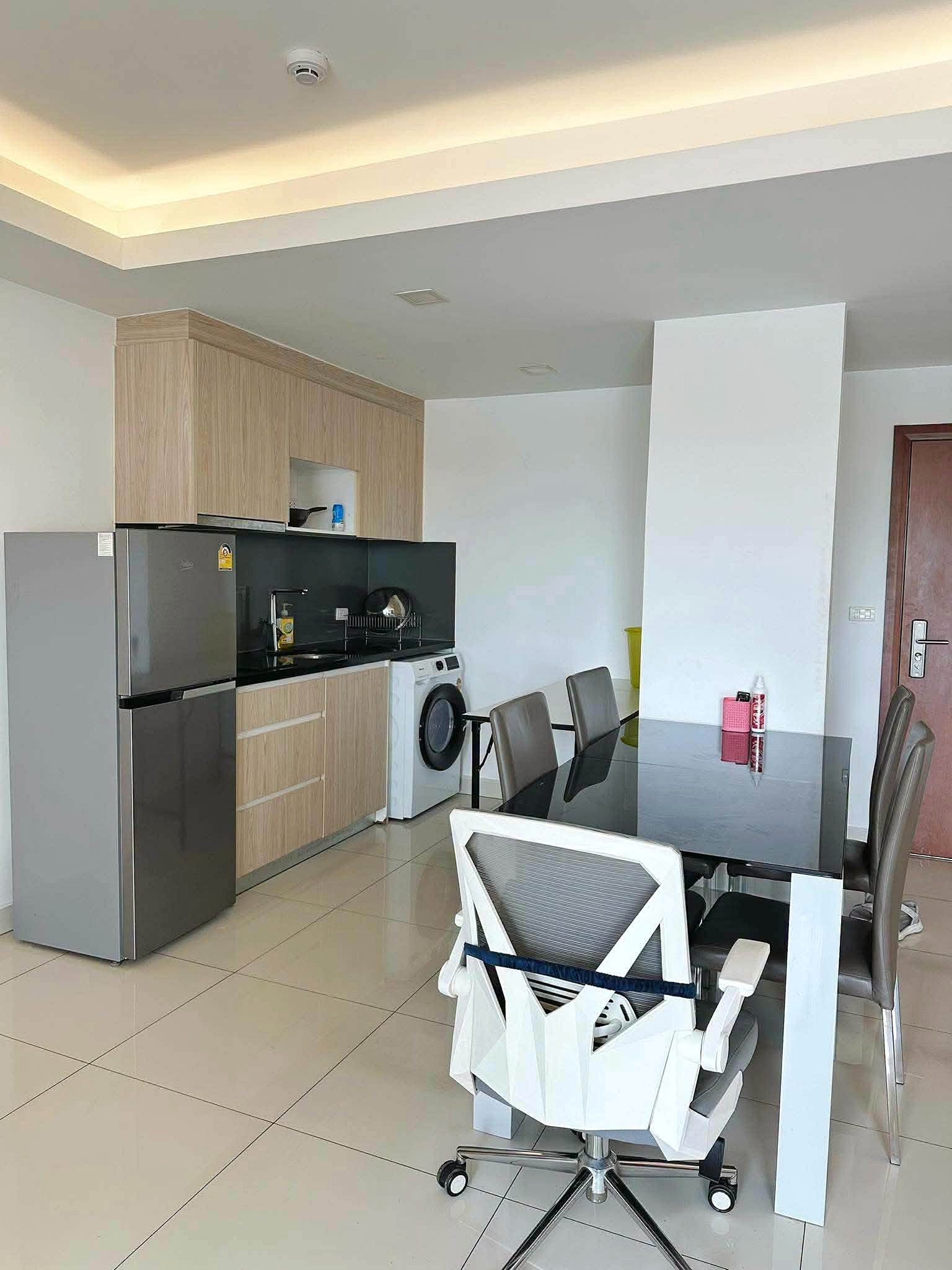Laguna_2_Pattaya_Sale_Dlc636_4 Modern kitchen and dining area with wooden cabinets, stainless steel fridge, washing machine, glass dining table, and gray chairs; white partition wall and tiled floor.