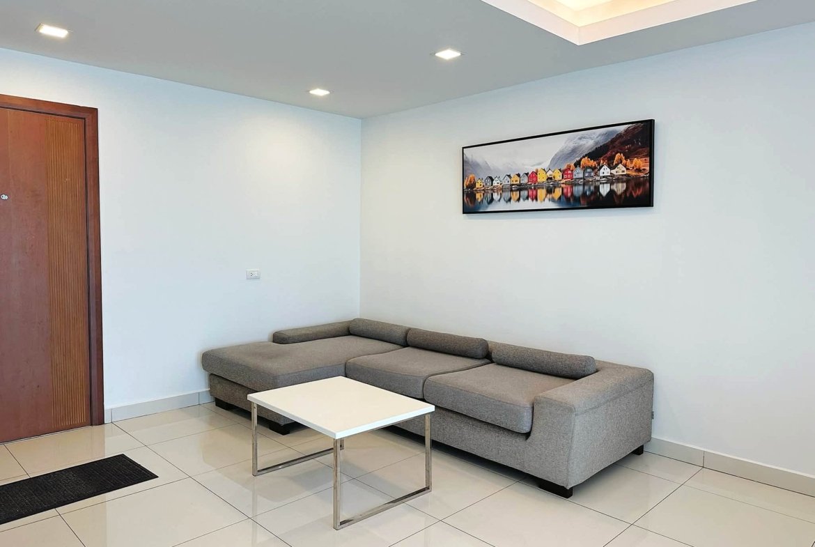 Modern living room corner with a gray L-shaped sofa, white coffee table, and a framed colorful village artwork on a light blue wall and tiled floor.