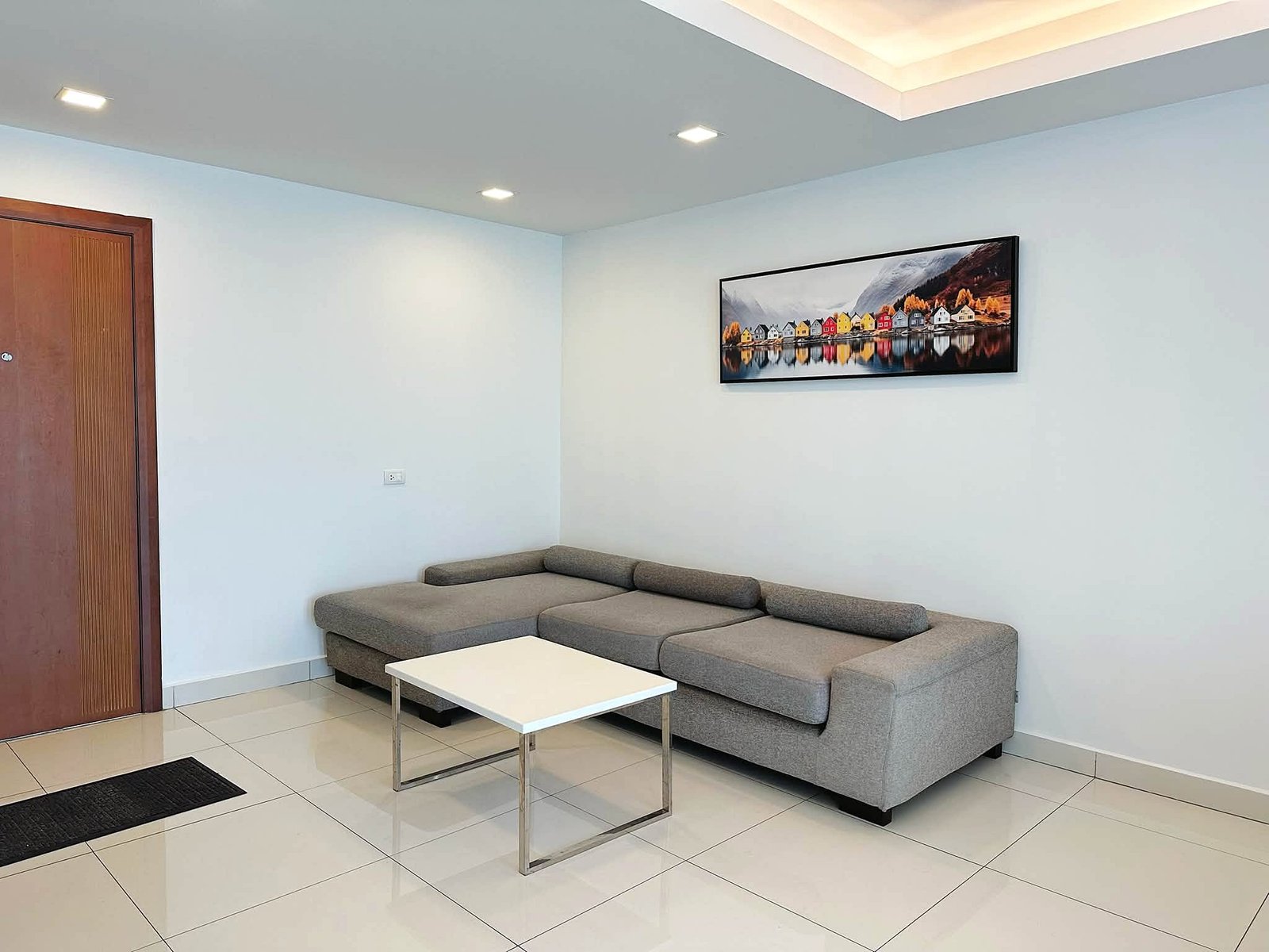 Laguna_2_Pattaya_Sale_Dlc636_5 Modern living room corner with a gray L-shaped sofa, white coffee table, and a framed colorful village artwork on a light blue wall and tiled floor.