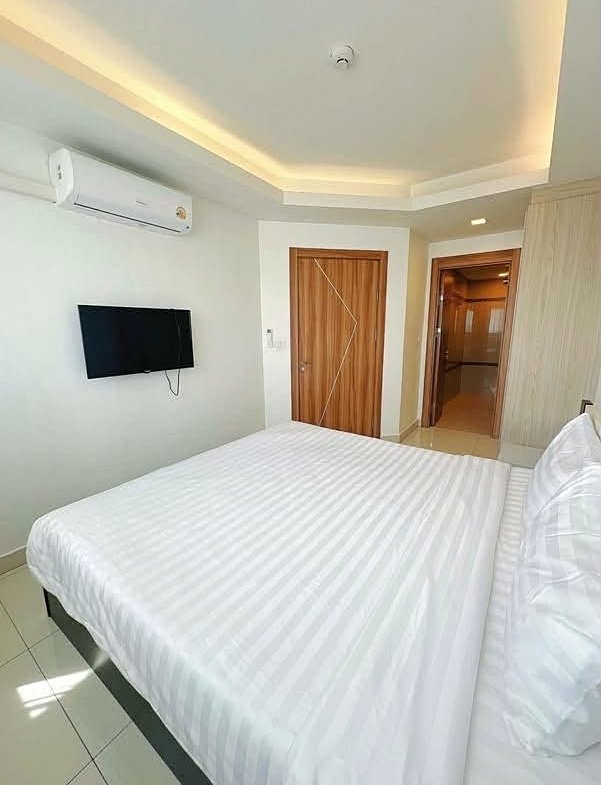 Hotel bedroom with a large bed in white striped linens, a wall-mounted flat-screen TV, and a wall air conditioner. A wooden door and a doorway to a hallway are visible in a bright, modern setting.