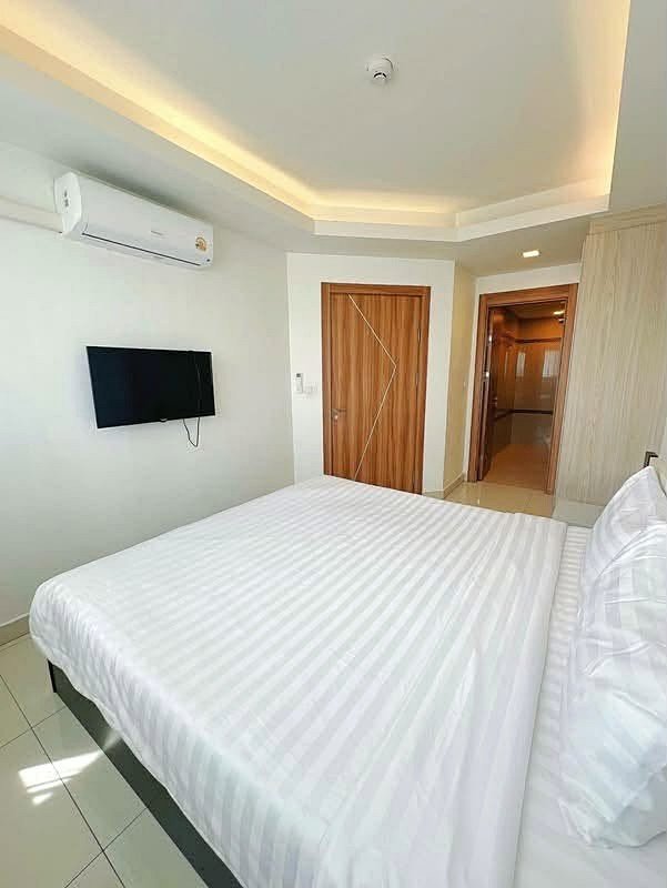 Laguna_2_Pattaya_Sale_Dlc636_6 Hotel bedroom with a large bed in white striped linens, a wall-mounted flat-screen TV, and a wall air conditioner. A wooden door and a doorway to a hallway are visible in a bright, modern setting.