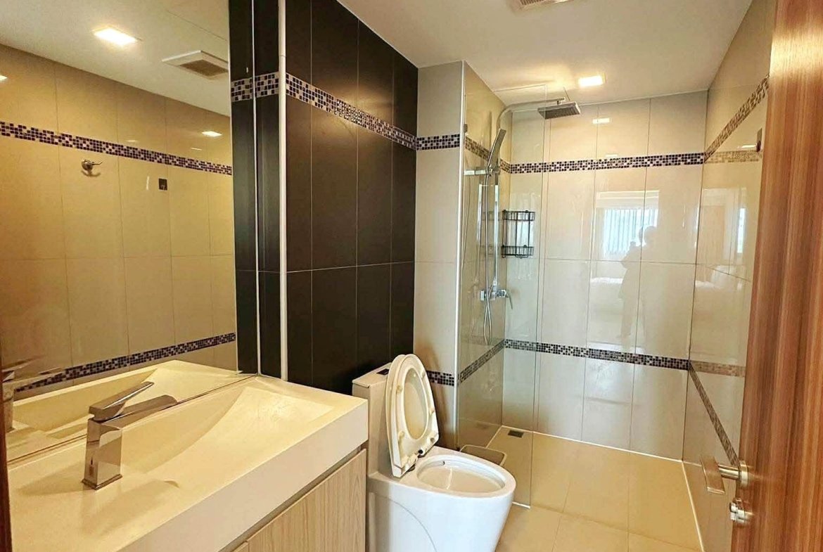 Modern bathroom with beige tiles, wooden vanity, and glass shower enclosure; toilet visible.