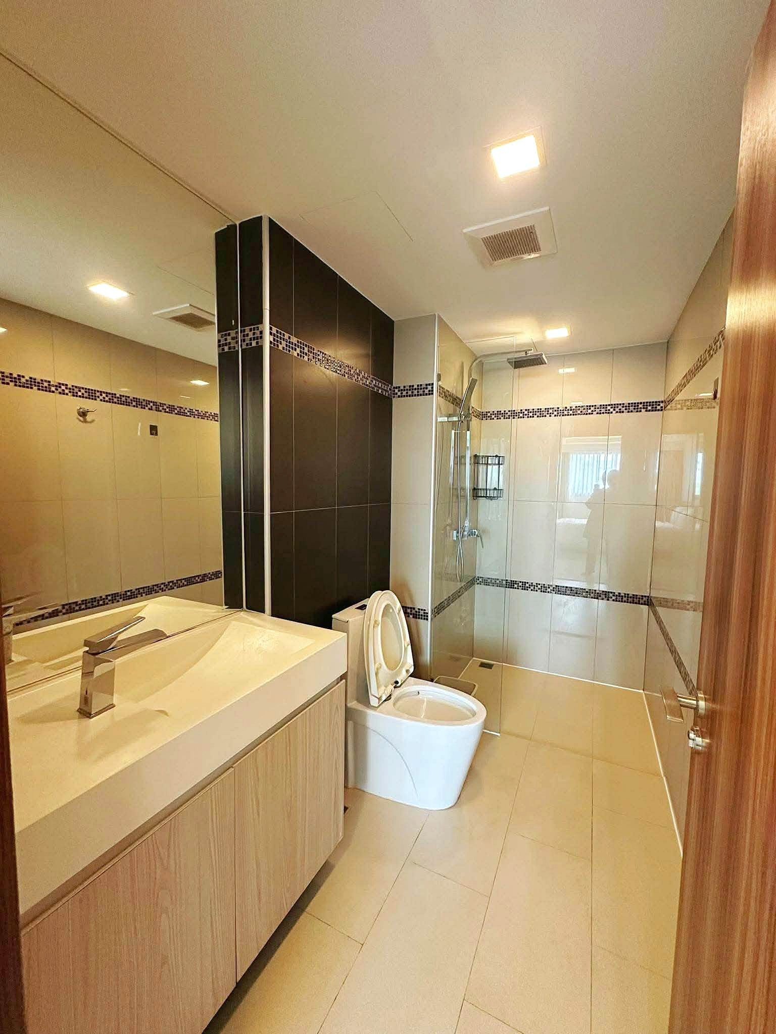Laguna_2_Pattaya_Sale_Dlc636_8 Modern bathroom with beige tiles, wooden vanity, and glass shower enclosure; toilet visible.