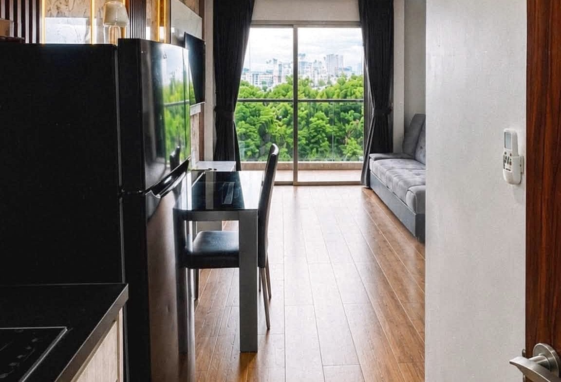 Open-plan kitchen and living area with a black refrigerator, glass dining table, and a balcony door showing green trees outside.