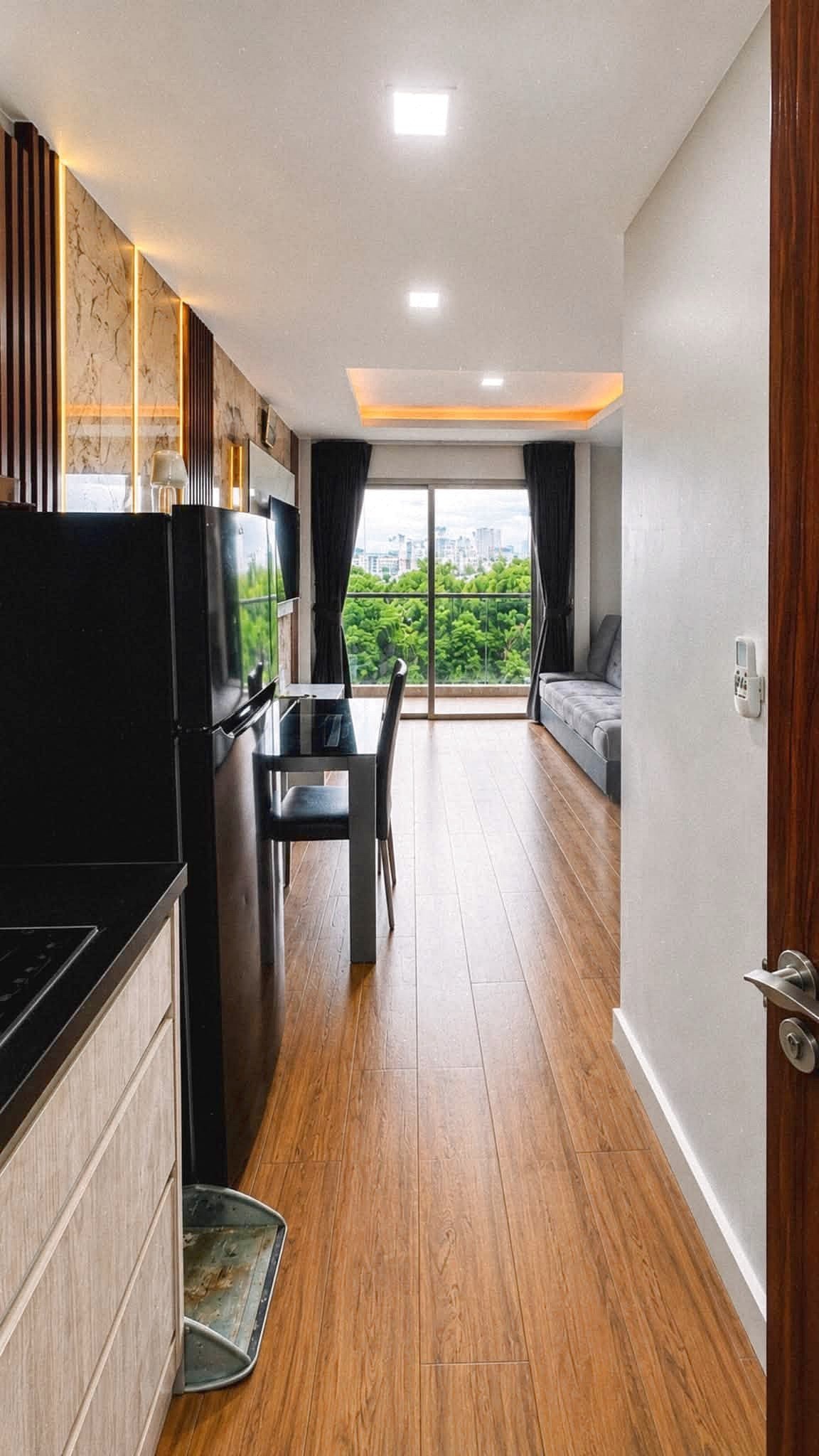 Laguna_2_Pattaya_Sale_Dlc644 Open-plan kitchen and living area with a black refrigerator, glass dining table, and a balcony door showing green trees outside.