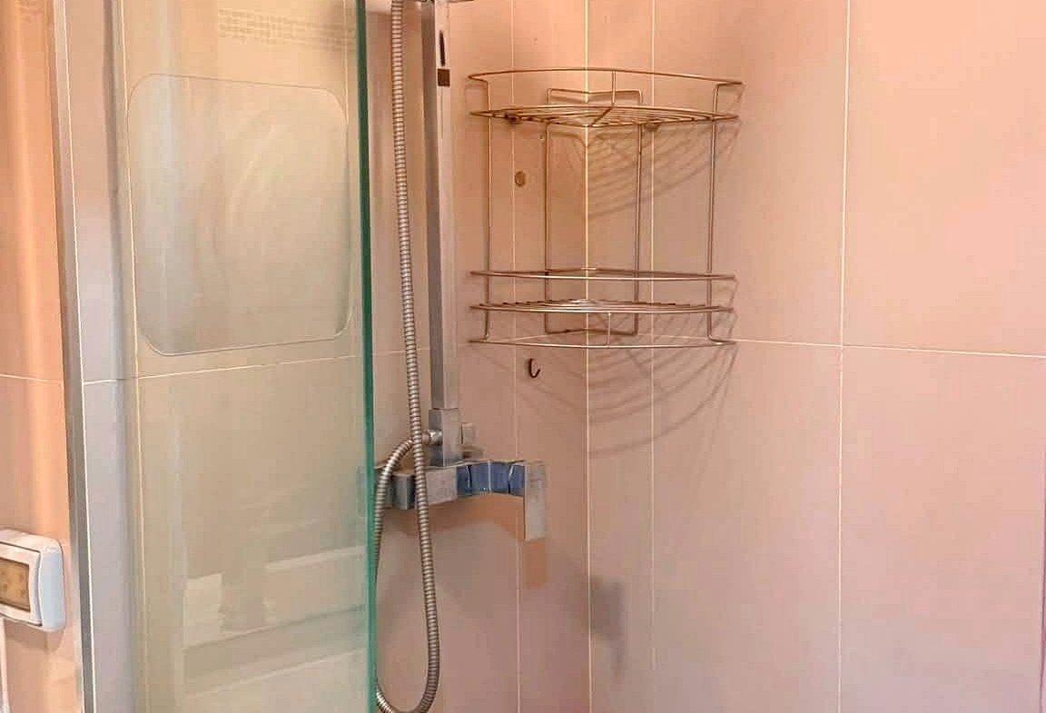 Glass-enclosed shower with a ceiling rainfall showerhead, handheld shower, and a chrome corner rack against tiled walls with mosaic stripe.