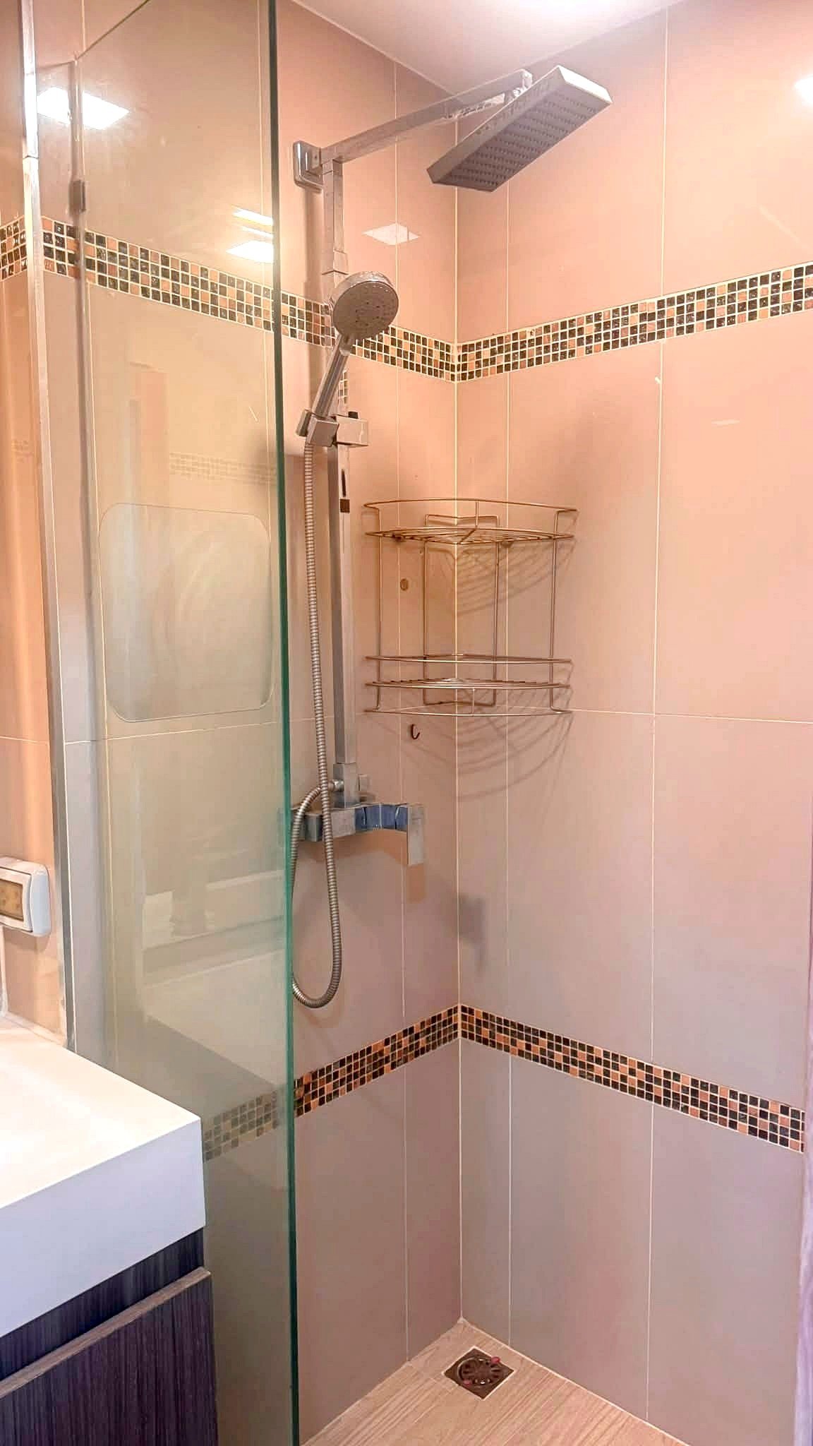Laguna_2_Pattaya_Sale_Dlc644_10 Glass-enclosed shower with a ceiling rainfall showerhead, handheld shower, and a chrome corner rack against tiled walls with mosaic stripe.