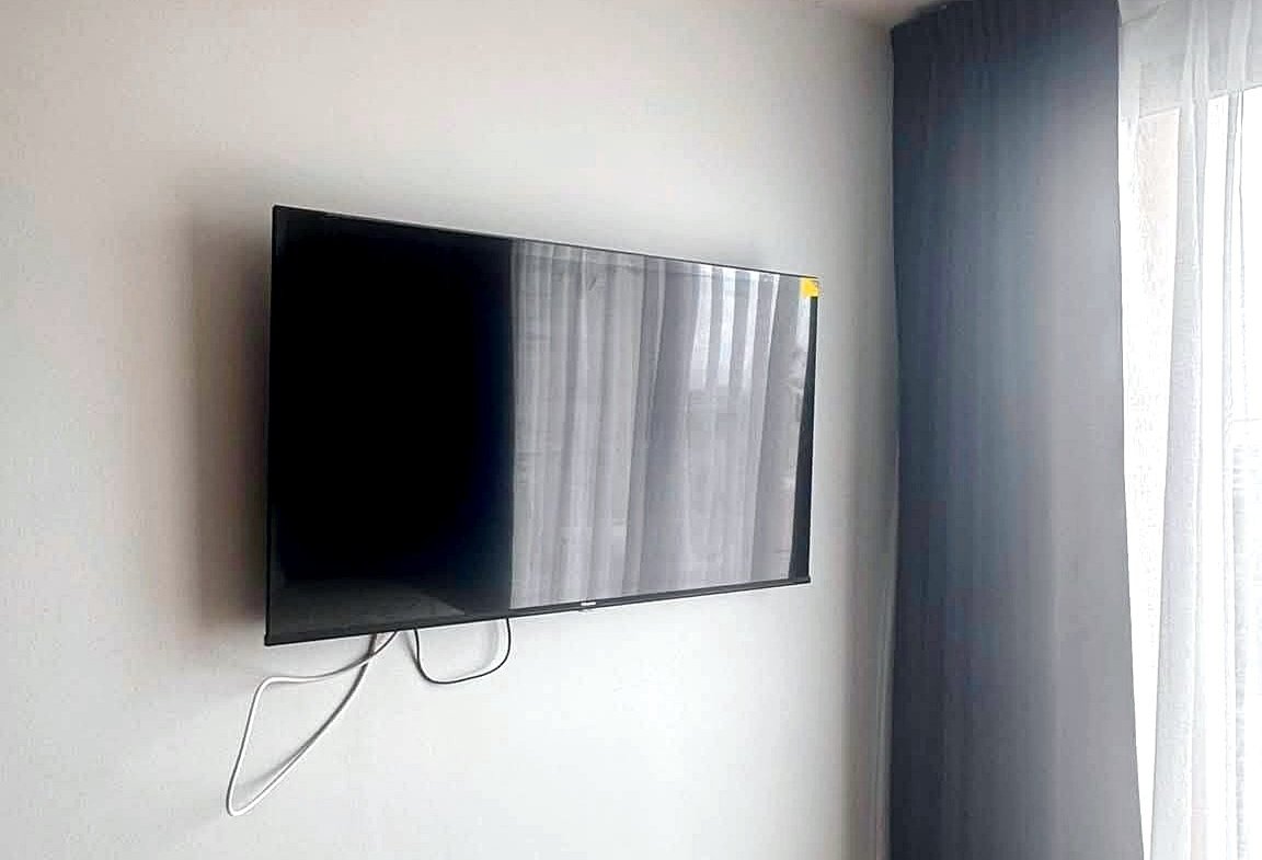 Wall-mounted flat-screen TV above a white geometric cabinet, remote on top and visible cables in a bright living space.