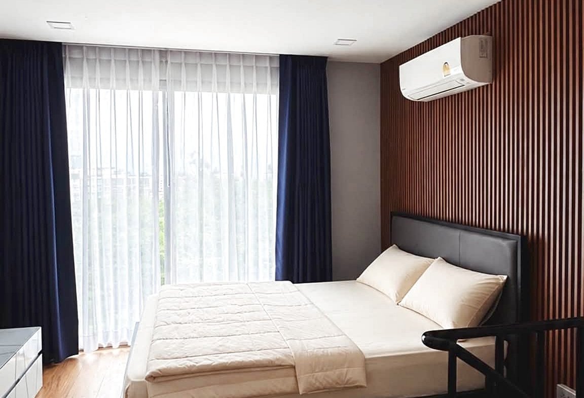Hotel bedroom with a low dark wood bed, beige linens, and a vertical wood slat accent wall; large window with sheer white curtains and navy drapes, wood floor, and wall-mounted AC unit.